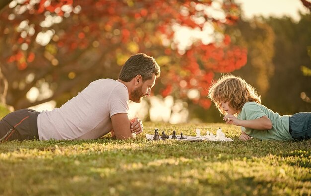 Familia feliz de papá e hijo jugando al ajedrez en la hierba verde en el parque erudito al aire libre