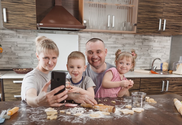 Familia feliz, papá, mamá y dos niños se toman selfies en la cocina mientras hacen galletas. cocinando juntos relaciones familiares felices