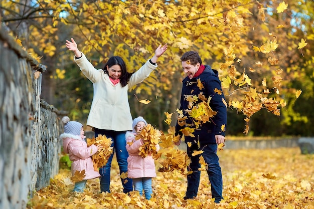 Familia feliz en el parque otoño. Madre, padre y dos niñas en la naturaleza tirando hojas amarillas, jugando, riendo.