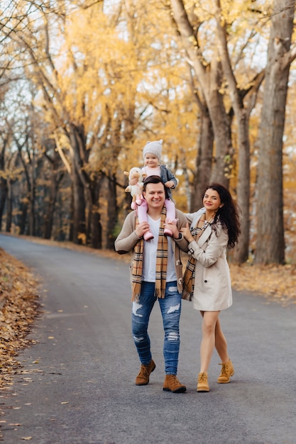 Familia feliz con el pequeño paseo del bebé en el camino del parque con los árboles amarillos en el otoño