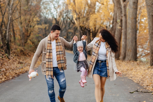 Familia feliz con el pequeño paseo del bebé en el camino del parque con los árboles amarillos en el otoño