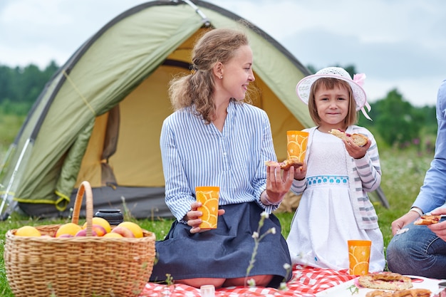 Familia feliz de picnic en el camping. Madre e hija comiendo cerca de una carpa en prado o parque