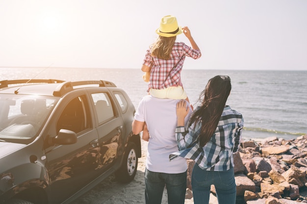 Familia feliz de pie cerca de un coche en la playa. Familia feliz en un viaje por carretera en su coche. Papá, mamá e hija viajan por el mar, el océano o el río. Paseo de verano en automóvil