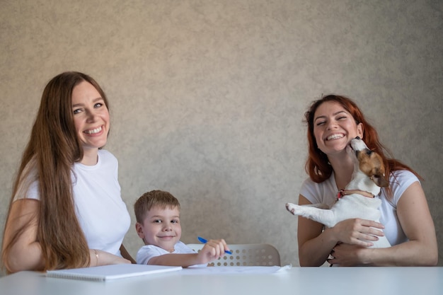 La familia feliz se queda en casa Dos mujeres ayudan al niño a hacer la tarea de la escuela Pareja de lesbianas sentadas a la mesa con su hijo y un cachorro alegre Matrimonio del mismo sexo con un niño Aprendizaje a distancia