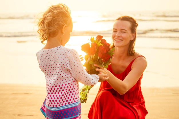 Familia feliz con ramo de flores rosas en la playa junto al mar al atardecer Joven hermosa asombrada madre e hija felicitando feliz cumpleaños Día de San Valentín 8 de marzo Día Internacional de la Mujer