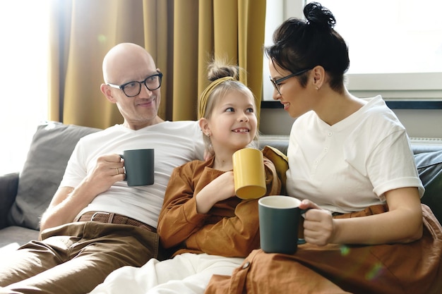 Familia feliz relajándose en el sofá y bebiendo té caliente en la sala de estar de su acogedor apartamento