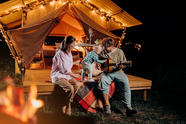 Família feliz relaxando e passando tempo juntos em glamping na noite de verão e tocando violão perto de uma fogueira aconchegante Barraca de acampamento de luxo para recreação ao ar livre e recreação Conceito de estilo de vida