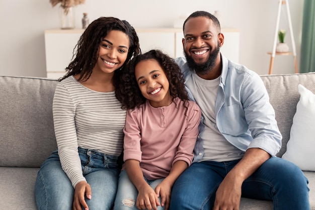 Familia feliz. Retrato de alegre hija afroamericana sentada en el sofá entre sus padres sonrientes. Mujer, hombre y niña positivos posando para la foto y mirando a la cámara en casa