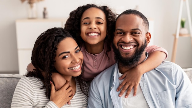 Família feliz. Retrato de alegre filha afro-americana, abraçando seus pais sorridentes por trás. Mulher positiva, homem e mulher posando para uma foto e olhando para a câmera em casa, sentados no sofá