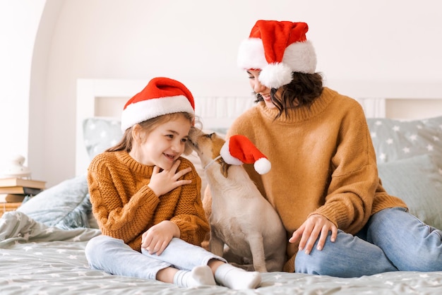 Família feliz se diverte com cachorro engraçado na cama em chapéus de papai noel Momentos emocionais