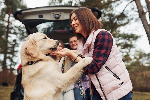 Família feliz se diverte com seu cachorro perto de carro moderno ao ar livre na floresta