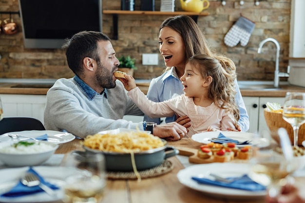 Família feliz se divertindo durante o almoço na mesa de jantar. A menina está alimentando seu pai.