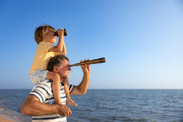 Família feliz se divertindo na praia avô e menino olhando através da luneta contra o fundo azul do mar e do céu férias de verão e conceito de dia dos pais