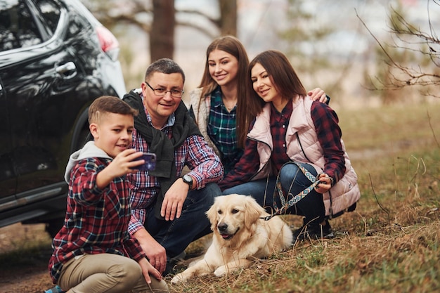 Familia feliz sentada y divirtiéndose con su perro cerca de un auto moderno al aire libre en el bosque