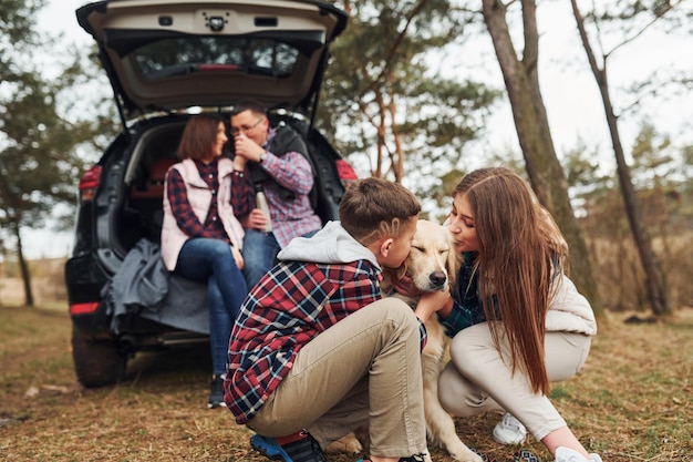 Familia feliz sentada y divirtiéndose con su perro cerca de un auto moderno al aire libre en el bosque