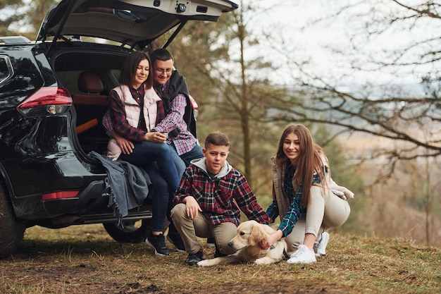 Familia feliz sentada y divirtiéndose con su perro cerca de un auto moderno al aire libre en el bosque