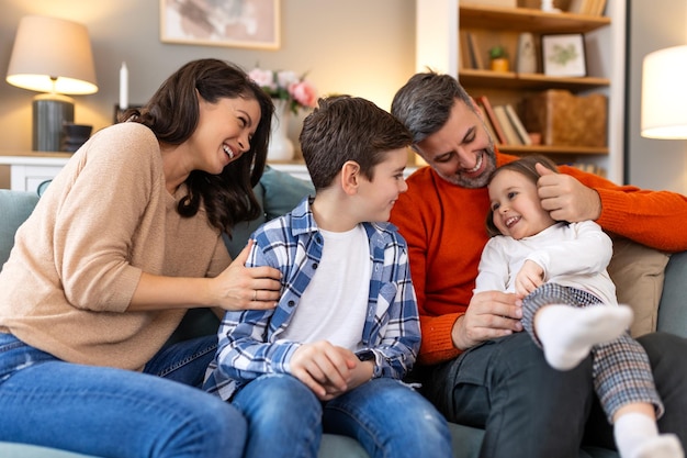 Foto familia feliz sentada en el sofá riendo juntos padres alegres jugando con sus hijos en casa el padre hace cosquillas a su hija pequeña mientras la madre y el hermano sonríen