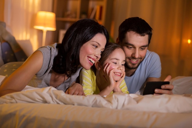 familia feliz con teléfono inteligente en la cama por la noche