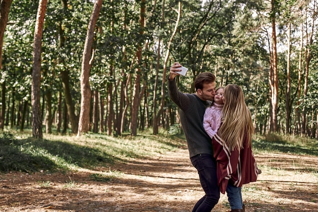 Familia feliz tomando selfie por teléfono inteligente en el bosque. Padre sostiene un teléfono inteligente con cámara