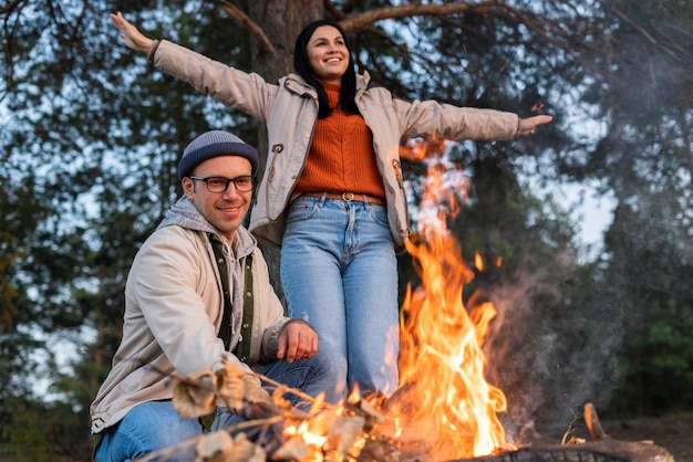 Familia feliz tomando el sol cerca del fuego en el acogedor bosque. Una mujer encantadora mirando hacia otro lado y extendiendo los brazos mientras su esposo se sentaba cerca