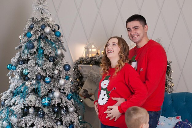 Familia feliz de tres madres jóvenes que esperan un nuevo bebé padre y su pequeño hijo cerca del árbol de Navidad decorado