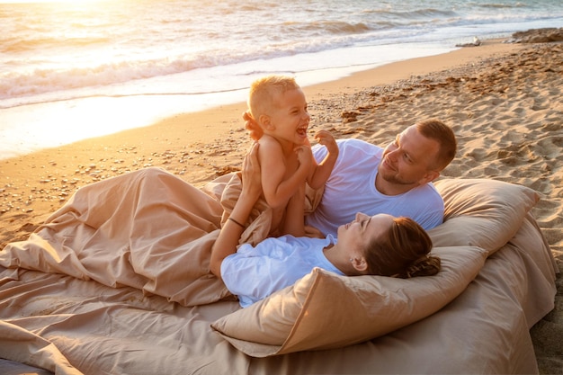 Una familia feliz de tres personas está descansando en una playa de arena mamá es una morena de pelo largo padre y ...