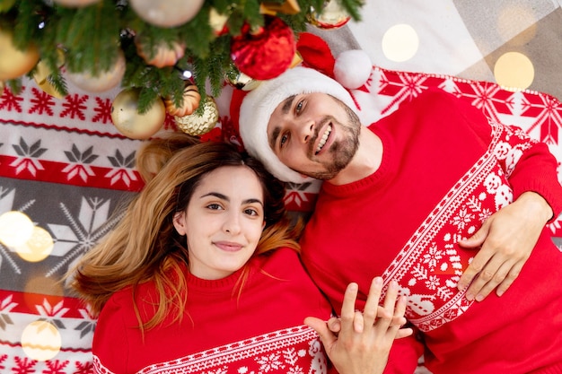 Família feliz um cara com uma garota de suéter vermelho sob a árvore de natal em casa comemora ou espera o ano novo ou o natal se alegrando e se abraçando