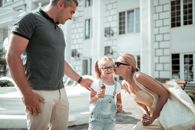 Família feliz. Uma menina segurando uma casquinha de sorvete enquanto a mãe a beijava e o pai segurava seu ombro durante a caminhada.