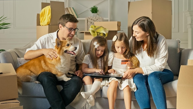 Una familia feliz usa una computadora portátil para comprar en línea, sentada en el sofá de su casa. Compra por Internet. Una familia sentada con un lindo perro en el sofá de casa. Confirmación de compra por Internet