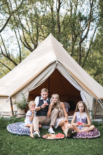 Familia feliz vistiendo ropa de vaquero boho, padre, madre y dos hijas sentados sobre almohadas en la hierba y comiendo sandía.