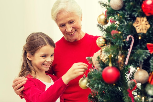 família, feriados, geração e conceito de pessoas - menina sorridente com a avó decorando a árvore de natal em casa