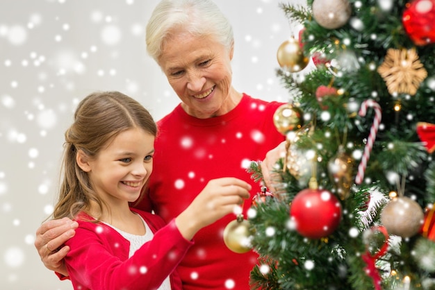 família, feriados, geração e conceito de pessoas - menina sorridente com a avó decorando a árvore de natal em casa