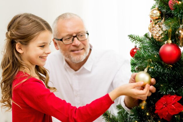 Família, feriados, geração e conceito de pessoas - menina sorridente com o avô decorando a árvore de natal em casa