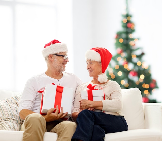 Família, feriados, idade e conceito de pessoas - casal sênior feliz em chapéus de ajudante de papai noel com caixas de presente sobre a sala de estar e o fundo da árvore de natal