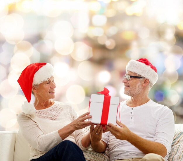 família, feriados, natal, idade e conceito de pessoas - casal sênior feliz em chapéus de ajudante de papai noel com caixa de presente sobre fundo de luzes