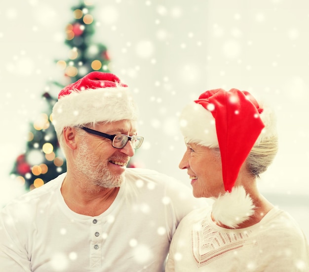família, feriados, natal, idade e conceito de pessoas - casal sênior feliz em chapéus de ajudante de papai noel olhando um para o outro em casa
