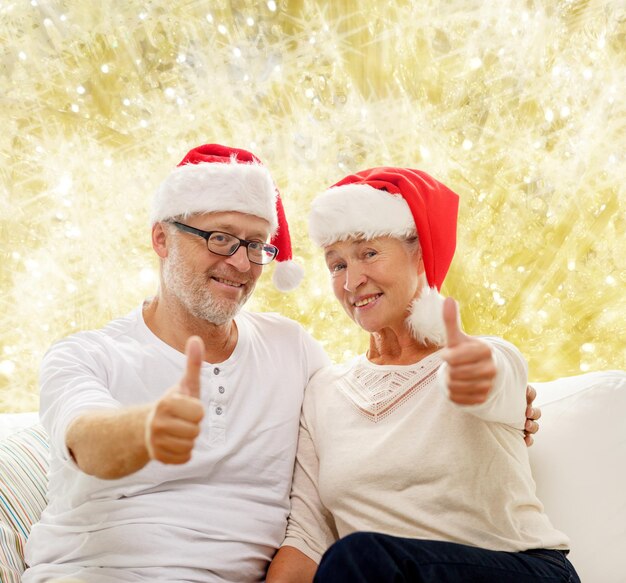 família, feriados, natal, idade e conceito de pessoas - casal sênior feliz em chapéus de ajudante de papai noel sentado no sofá sobre fundo de luzes amarelas e mostrando os polegares para cima gesto