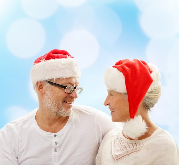 Família, feriados, natal, idade e conceito de pessoas - casal sênior feliz em chapéus de ajudante de papai noel sentado no sofá sobre fundo de luzes azuis