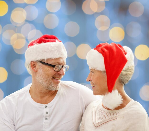 família, feriados, natal, idade e conceito de pessoas - casal sênior feliz em chapéus de ajudante de papai noel sentado no sofá sobre fundo de luzes azuis