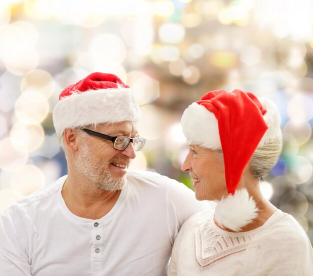 família, feriados, natal, idade e conceito de pessoas - casal sênior feliz em chapéus de ajudante de papai noel sentado no sofá sobre fundo de luzes