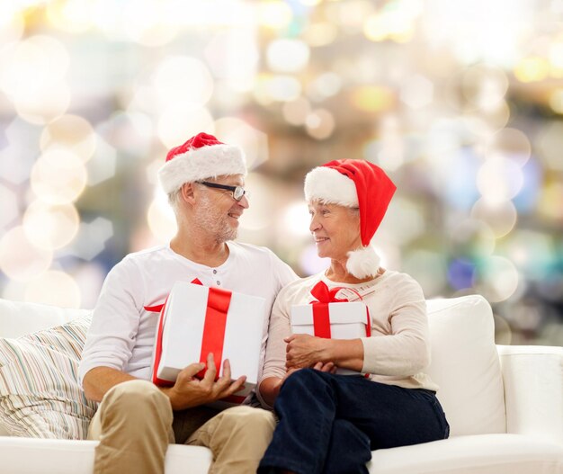 família, feriados, natal, idade e conceito de pessoas - feliz casal sênior em chapéus de ajudante de papai noel com caixas de presente sobre fundo de luzes