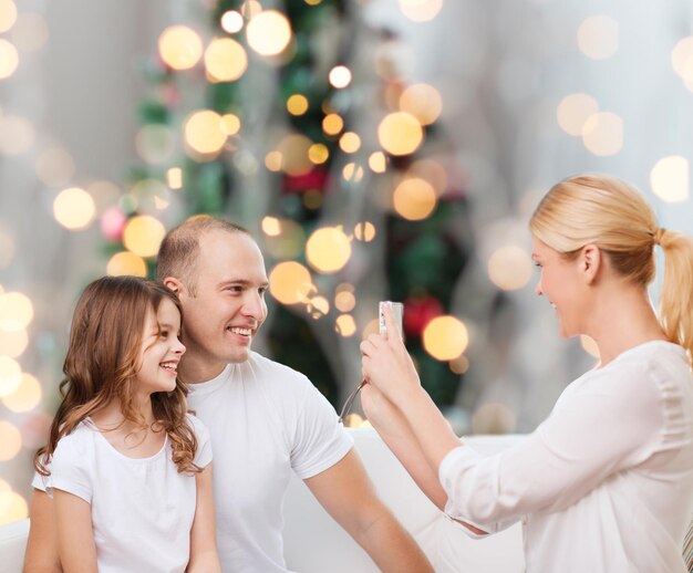 família, feriados, tecnologia e conceito de pessoas - sorrindo mãe, pai e filha com câmera sobre fundo de luzes da árvore de natal