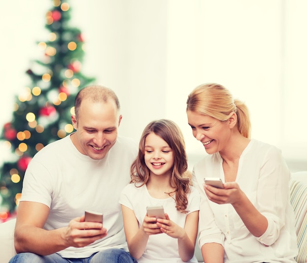 família, feriados, tecnologia e pessoas - sorrindo mãe, pai e filha com smartphones sobre a sala de estar e o fundo da árvore de natal