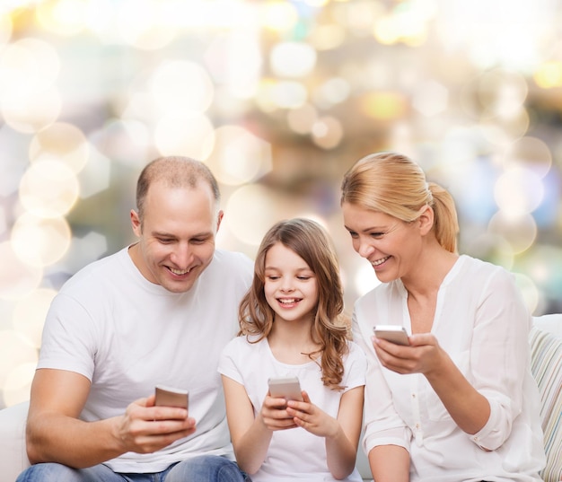 família, feriados, tecnologia e pessoas - sorrindo mãe, pai e filha com smartphones sobre fundo de luzes