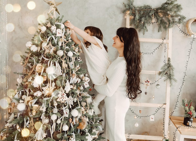 Família, férias de inverno e conceito de pessoas - mãe feliz segura a filha nos braços e decora a árvore de Natal em casa