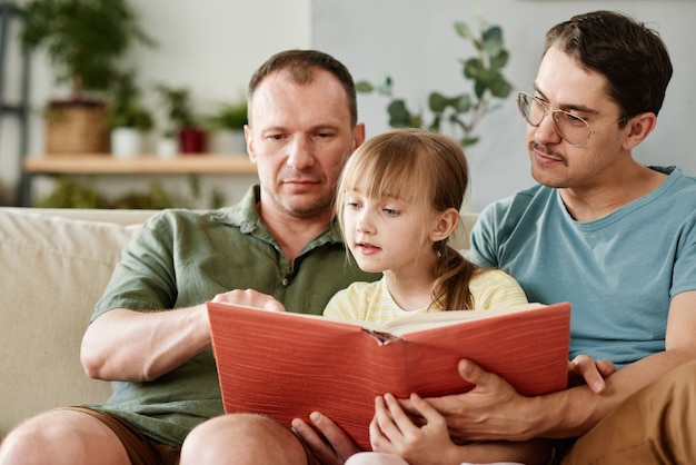 Família gay ensinando menina a ler um livro sentada no sofá da sala