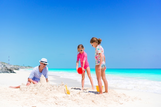 Familia haciendo castillos de arena en la playa blanca tropical, padre y dos niñas jugando con arena en la playa tropical