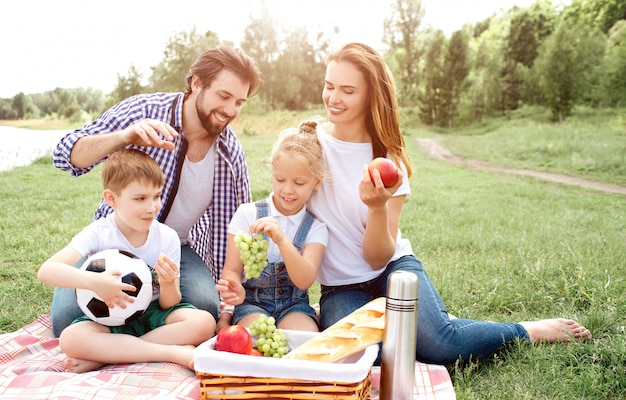 Familia hambrienta está sentado sobre una manta y mirando la canasta con comida. La mujer tiene manzana en sus manos. Chica tiene uvas en las manos. Niño está comiendo un trozo de uva. El hombre quiere agarrar comida de la canasta