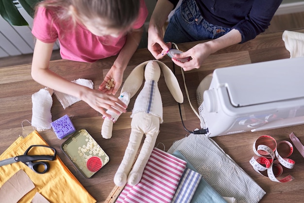 Família hobby e lazer criativos feitos à mão, crianças juntas sentadas em casa, costurando a boneca coelhinha de brinquedo. Garotas riem, aprendem técnicas de costura