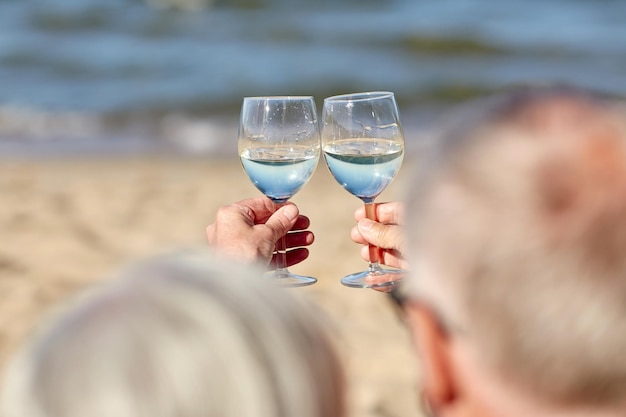 família, idade, férias, lazer e conceito de pessoas - close-up de casal sênior feliz tilintando copos de vinho na praia de verão